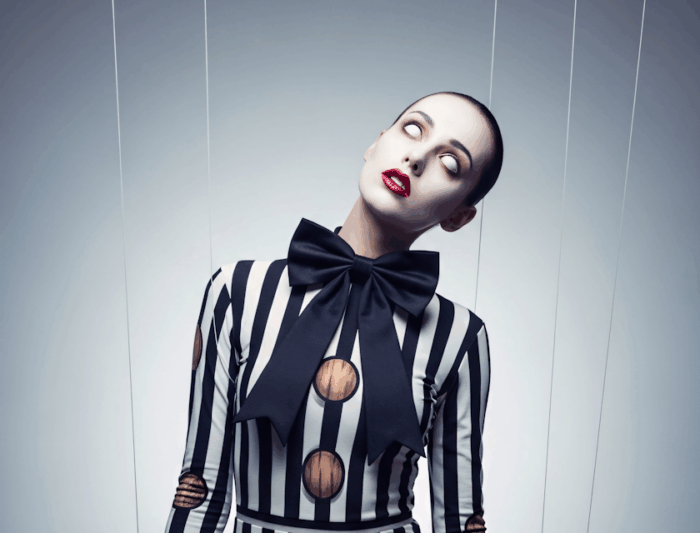 A person dressed as a marionette puppet, wearing a black and white vertically striped bodysuit with large wooden buttons and a giant black bow at the collar. Their face is painted white with exaggerated red lips and rolled-back eyes, and they are suspended by visible strings, mimicking a puppet pose.